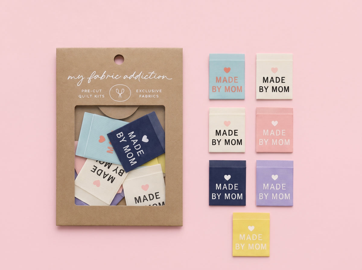 A kraft paper package labeled 'my fabric addiction' with a clear window showing folded fabric swatches, alongside six loose fabric swatches in pastel blue, cream, pink, navy, lavender, and yellow, each printed with the words 'MADE BY MOM' on a soft pink background.
