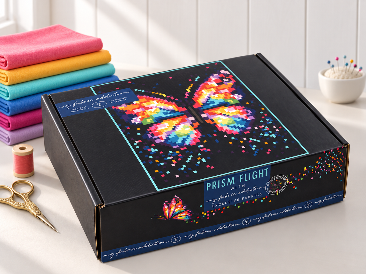 A black product box labeled 'PRISM FLIGHT' with a colorful butterfly graphic on top, styled on a white tabletop with folded fabric swatches, thread, scissors, and a pin cushion in the background.