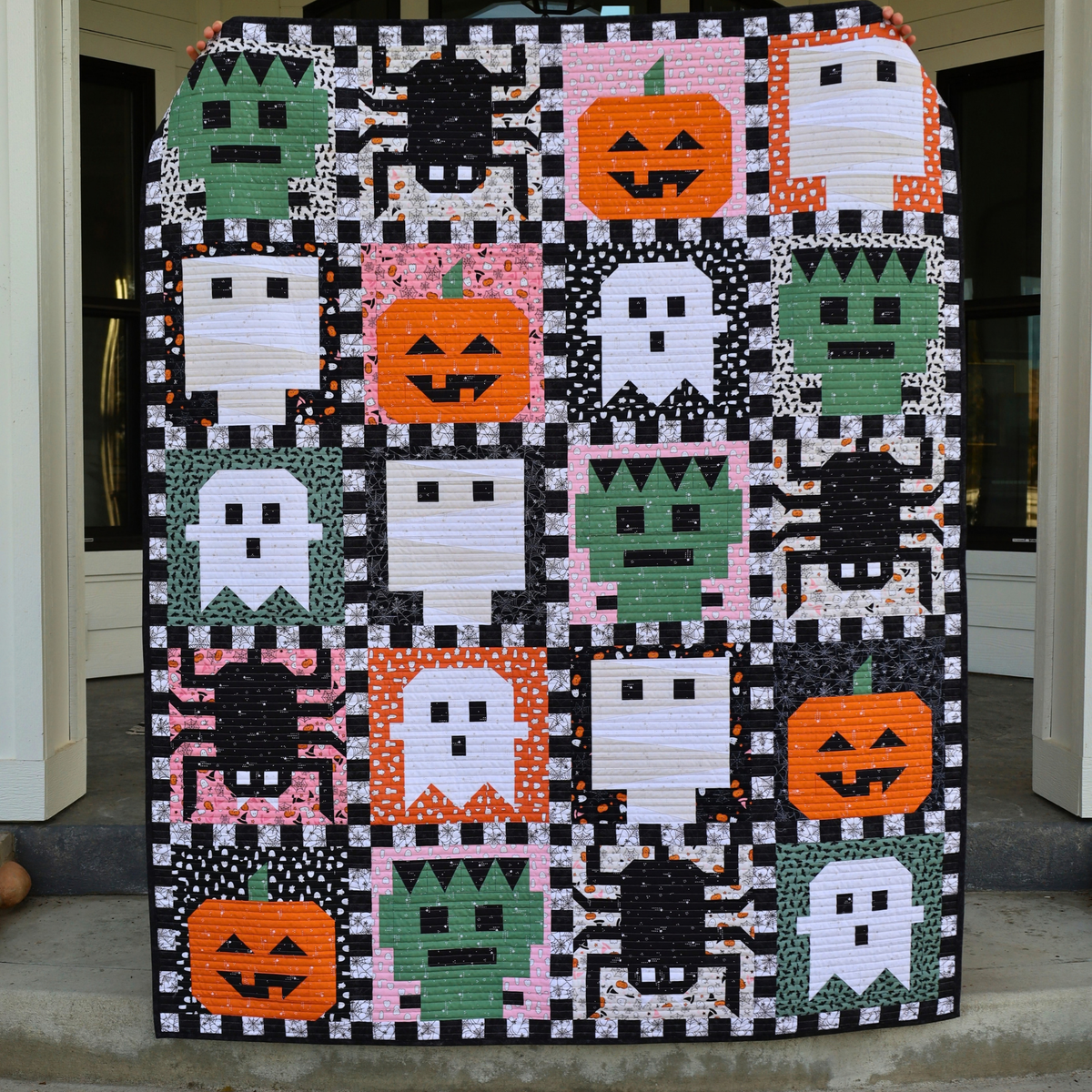 A handmade quilt featuring a grid of Halloween-themed patchwork squares with pumpkins, ghosts, spiders, and Frankenstein's monster on a black-and-white checkered background, displayed on a front porch.