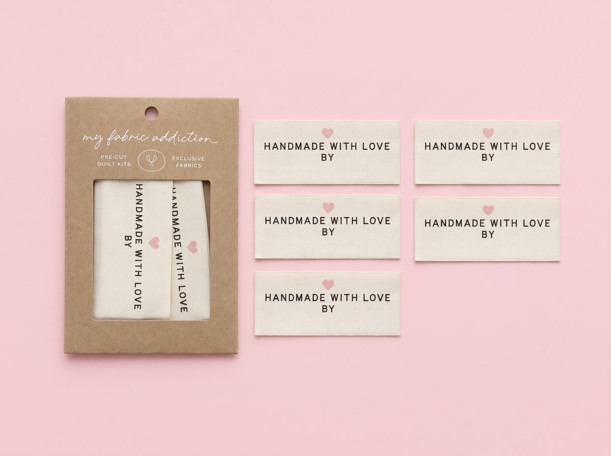 A set of cream fabric labels with black text reading 'HANDMADE WITH LOVE BY' and small pink hearts, shown next to their kraft paper packaging on a solid pink background.