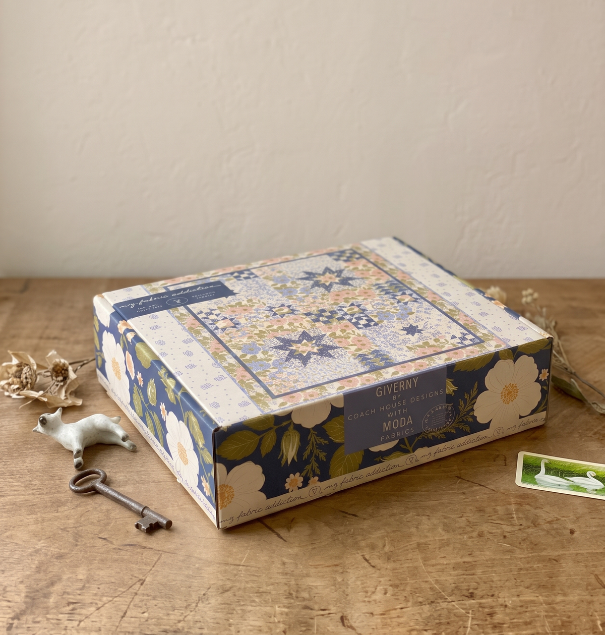 A decorative product box with a floral quilt pattern lid sits on a wooden tabletop against a beige wall. The box is labeled 'BISHOP' and 'SILKY CRAFTS', with small props including a vintage key, a small white dog figurine, and dried flowers around it.
