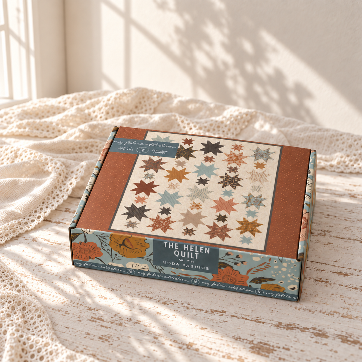 A quilt kit box labeled 'The Helen Quilt' sits on a cream lace blanket in warm sunlight. The box features a quilt pattern illustration with star and diamond shapes in muted orange, brown, blue, and cream tones, with 'My Fabric Collection' branding visible.