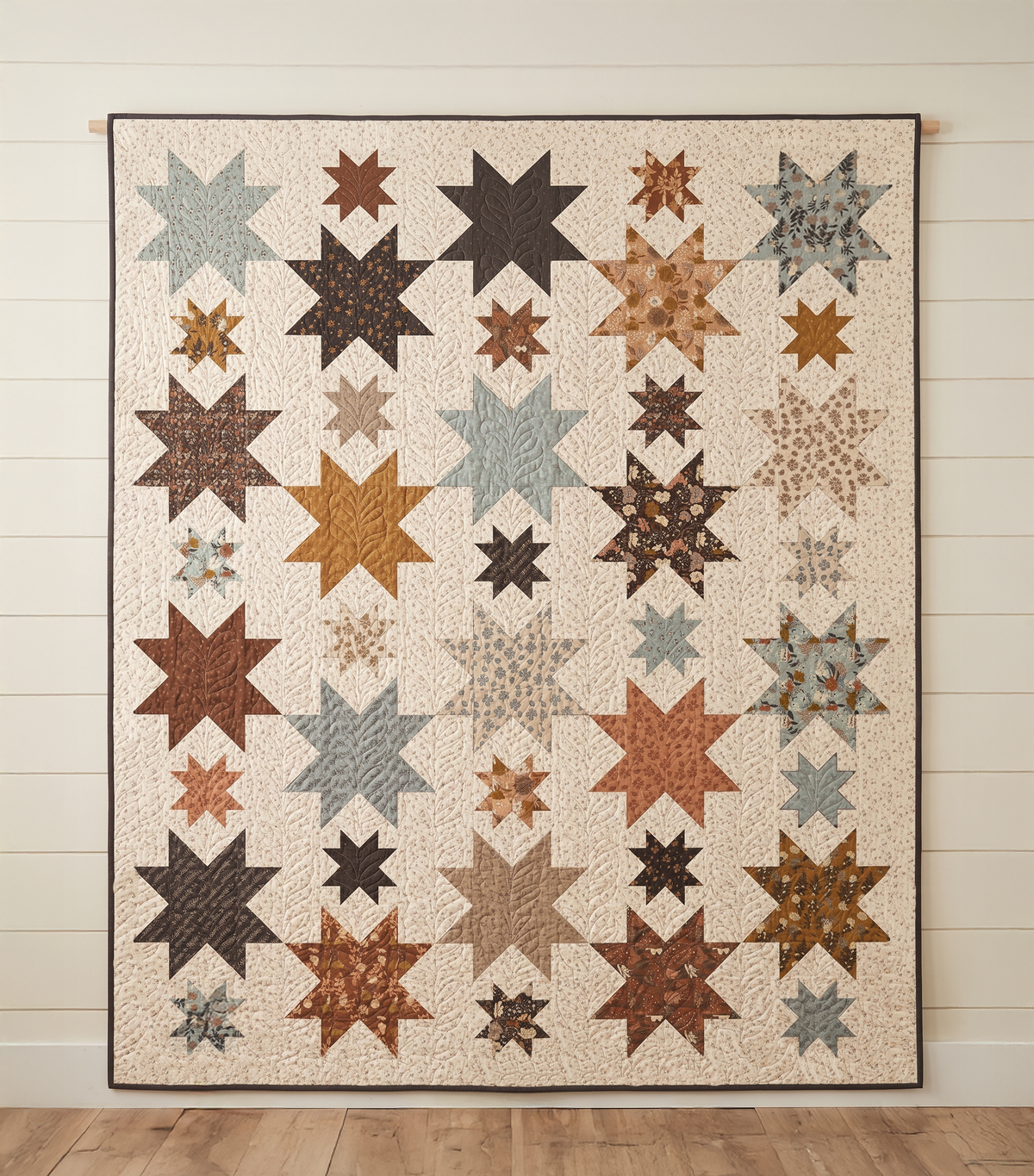 A quilted wall hanging featuring a grid of star and snowflake-like starburst motifs in earthy tones, including brown, beige, gray, and rust orange, displayed against a white shiplap wall.