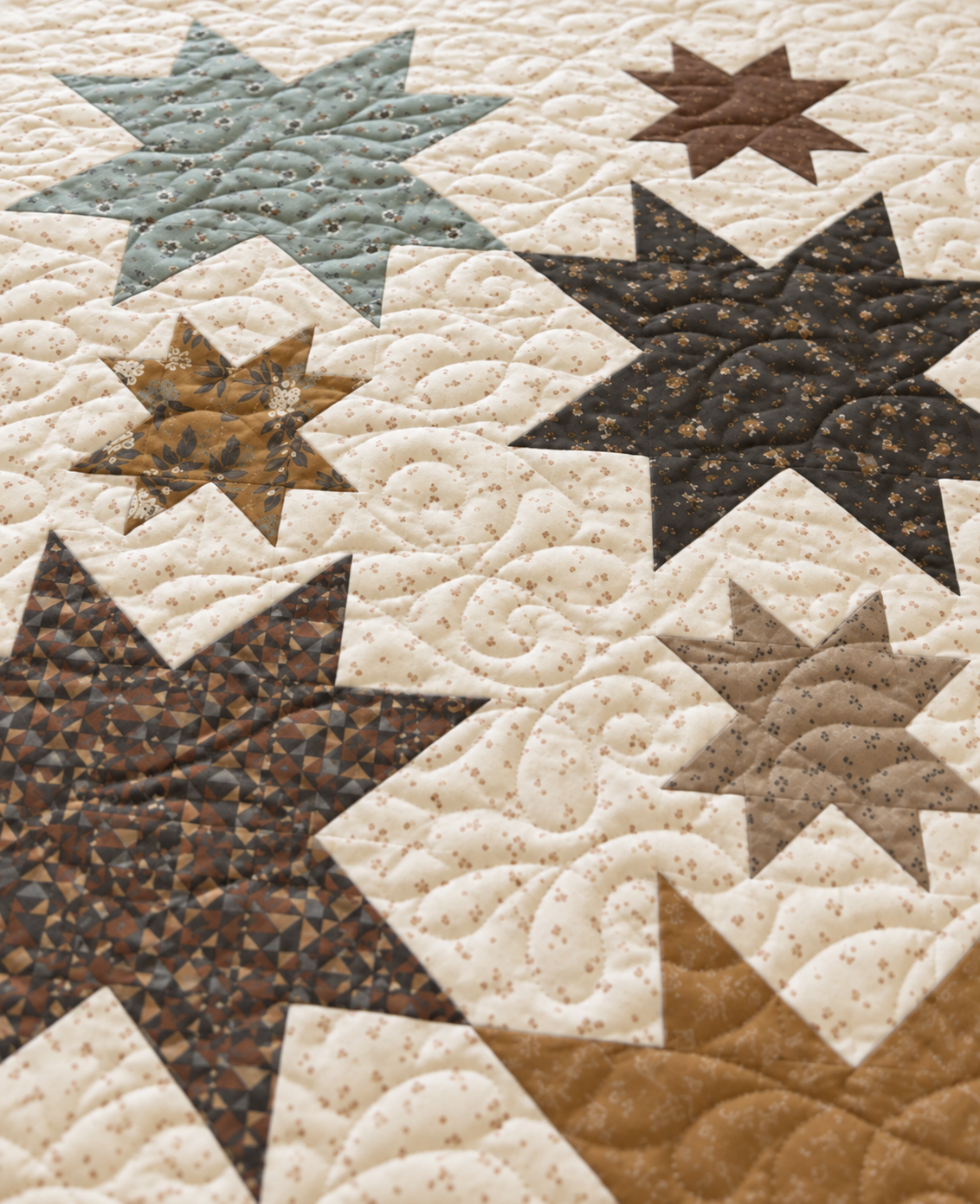 Close-up of a quilted fabric surface with large star and geometric patchwork motifs in cream, taupe, brown, and muted blue-gray, showing stitched quilting patterns.