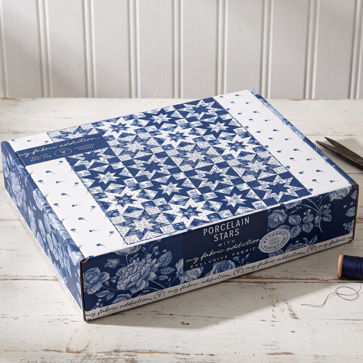 A fabric box by Porcelain Stars with a blue and white geometric and floral pattern, displayed on a white wooden table next to scissors and spools of thread.