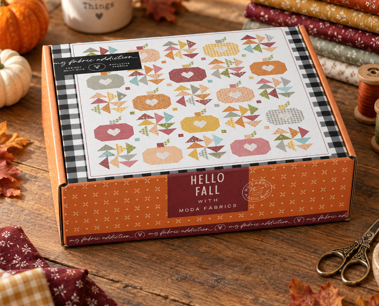 A boxed quilting fabric collection labeled 'HELLO FALL with MOOA FABBICS' sits on a wooden table surrounded by autumn-themed props, including fabric rolls, thread spools, leaves, and a small pumpkin. The box lid shows a colorful quilt pattern with pumpkins, hearts, and geometric motifs.