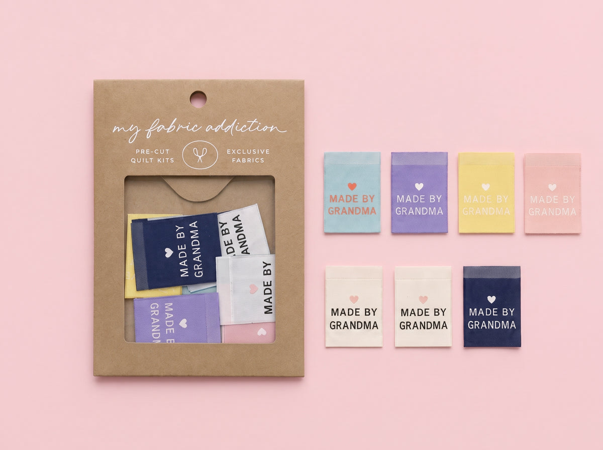 A kraft paper package labeled 'my fabric addiction' with a clear window showing folded fabric swatches, placed beside eight small fabric swatches in pastel and navy colors printed with 'MADE BY GRANDMA' and 'GRANDMA' in different colorways, all on a soft pink background.