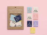 A kraft paper package labeled 'my fabric addiction' with a clear window showing folded fabric swatches, alongside six loose fabric swatches in pastel blue, cream, pink, navy, lavender, and yellow, each printed with the words 'MADE BY MOM' on a soft pink background.