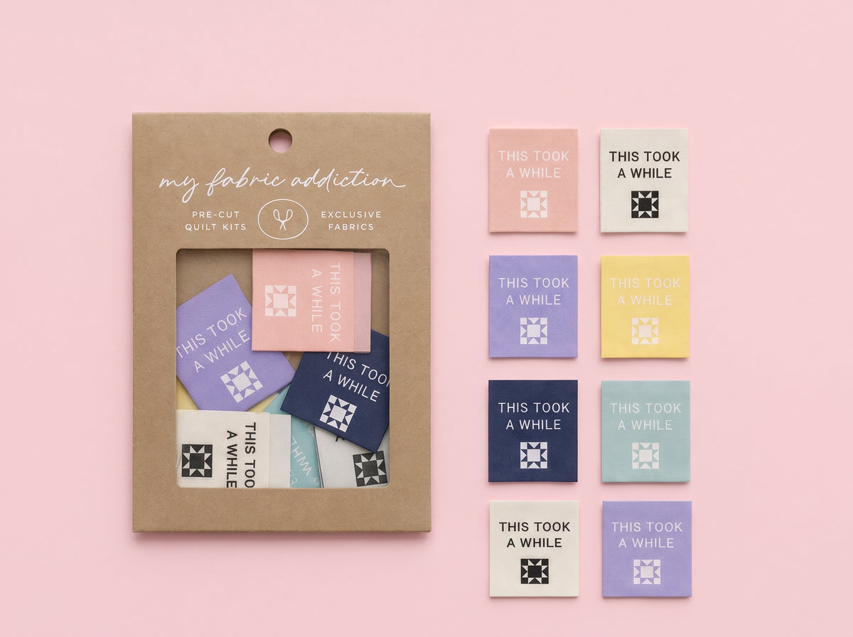 My Fabric Addiction pre-cut quilt mix in a kraft paper window box, displayed next to a grid of ten sample fabric swatches in pastel and dark blue shades on a solid pink background.